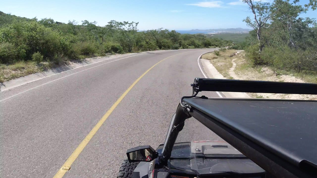 Forward view from a Wrangler on a curving empty road in Los Cabos, Mexico, with green brush, pale shoulder, distant hills, and no visible people.