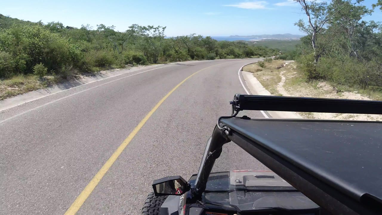 Forward-facing vehicle view as the Los Cabos road curves left, with the Wrangler hardware low in frame, roadside scrub, distant mountains, and no visible people.