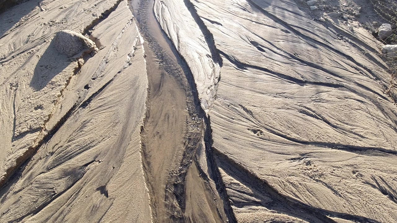 Opening overhead frame of pale sand and smooth stone in Los Cabos, Mexico, with a narrow water channel cutting through the center and no visible people.