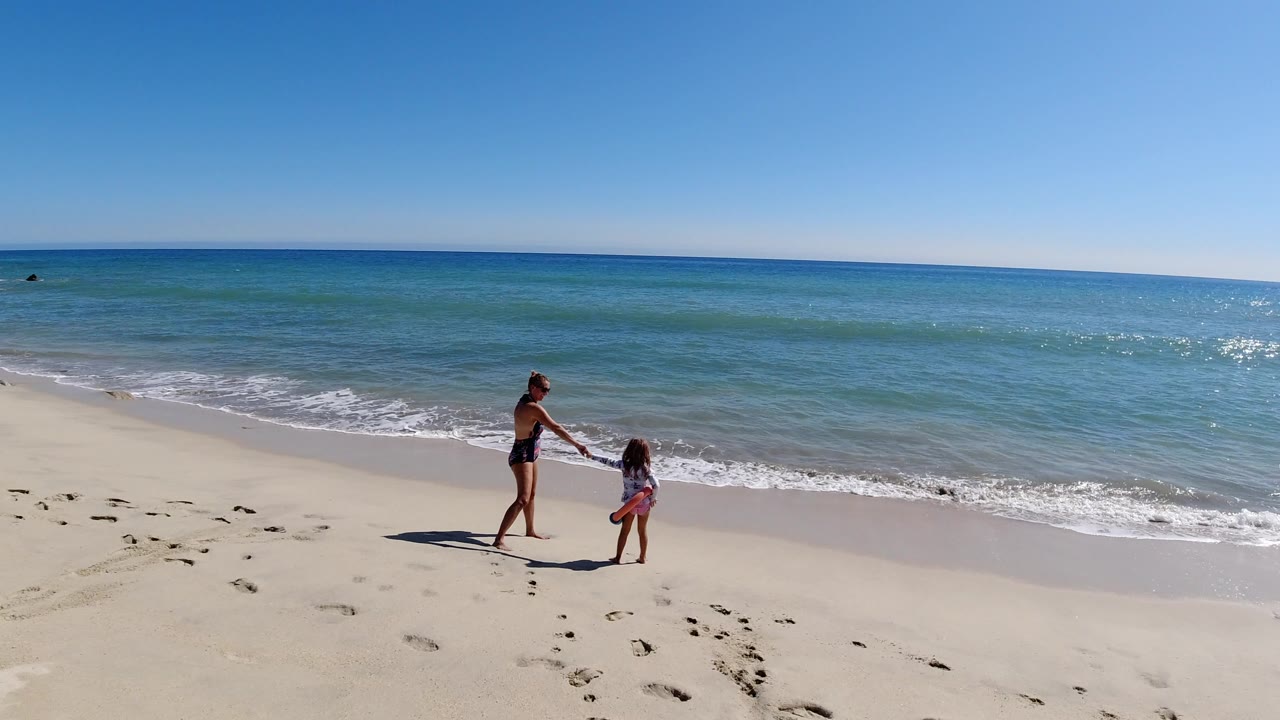 In Los Cabos, Mexico, a woman and young child hold hands on pale sand near small incoming waves, with blue-green ocean and a clear horizon behind them.