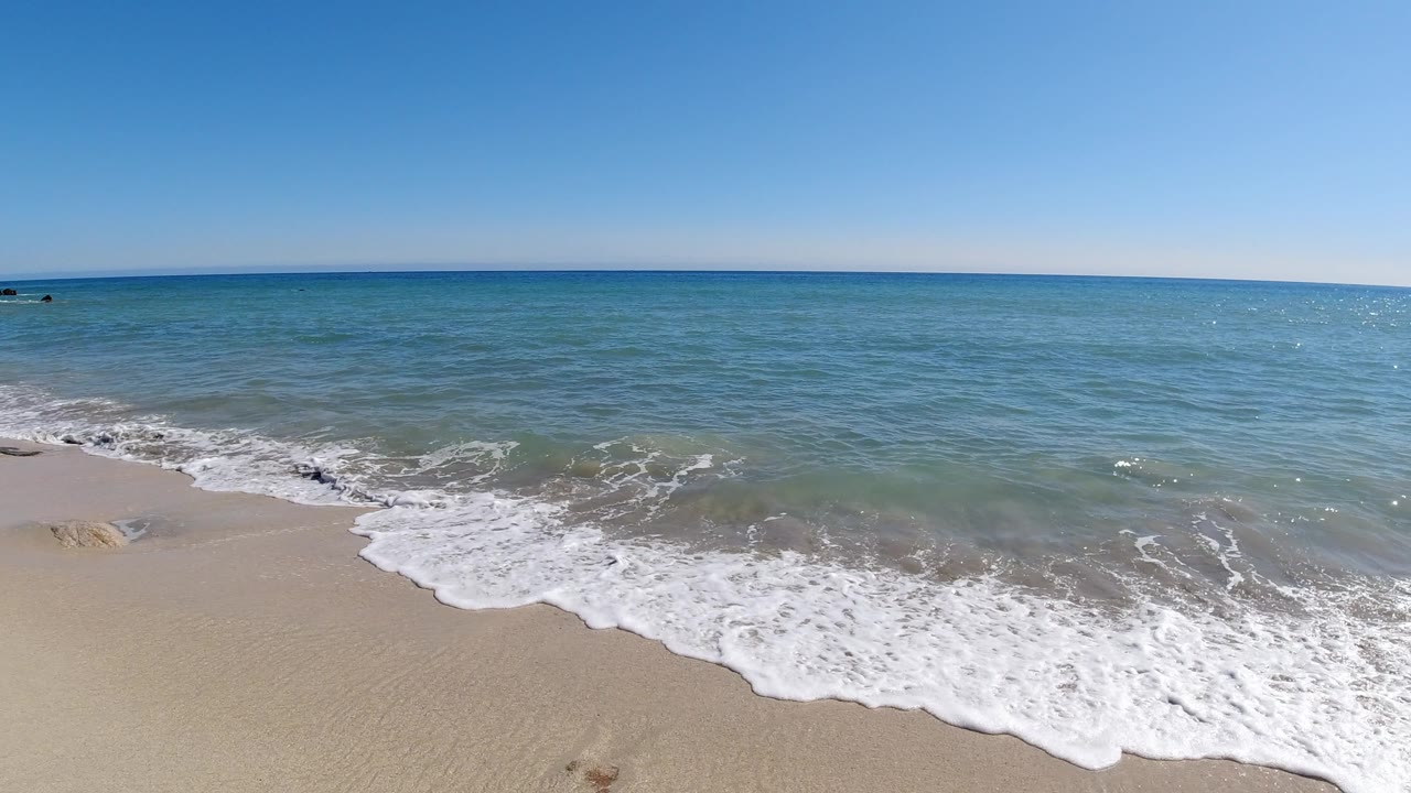A wider sheet of white foam rolls up the Los Cabos beach, with shallow green-blue water behind it, open sea at the horizon, and no visible people.