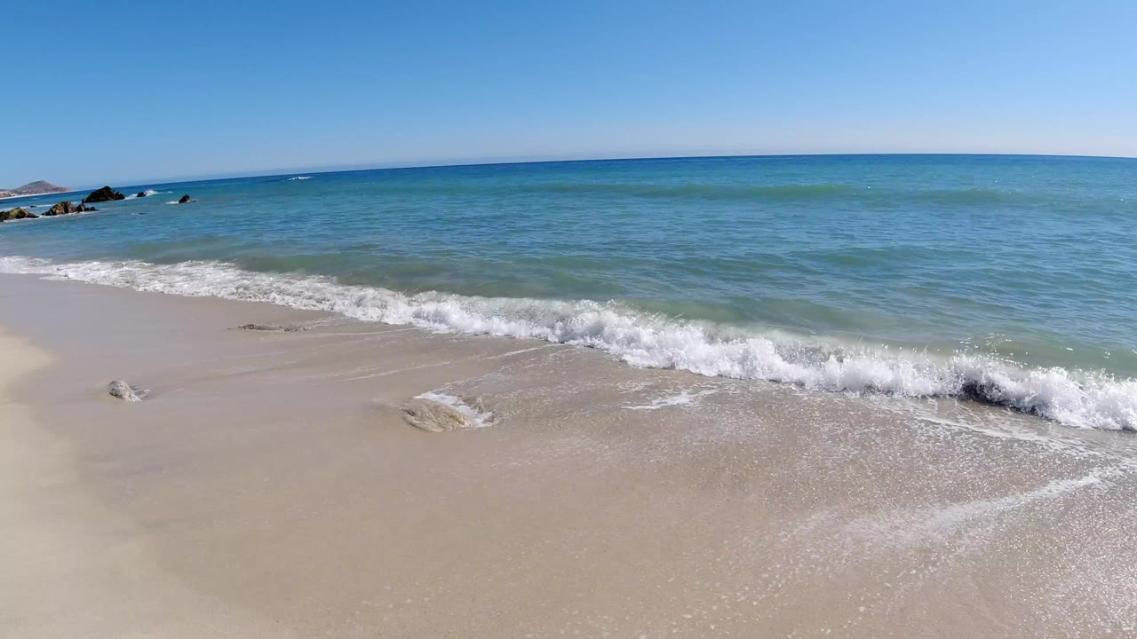 Los Cabos white sand beach at the start of the clip, with a small wave breaking near the foreground, blue-green water beyond, and no visible people.