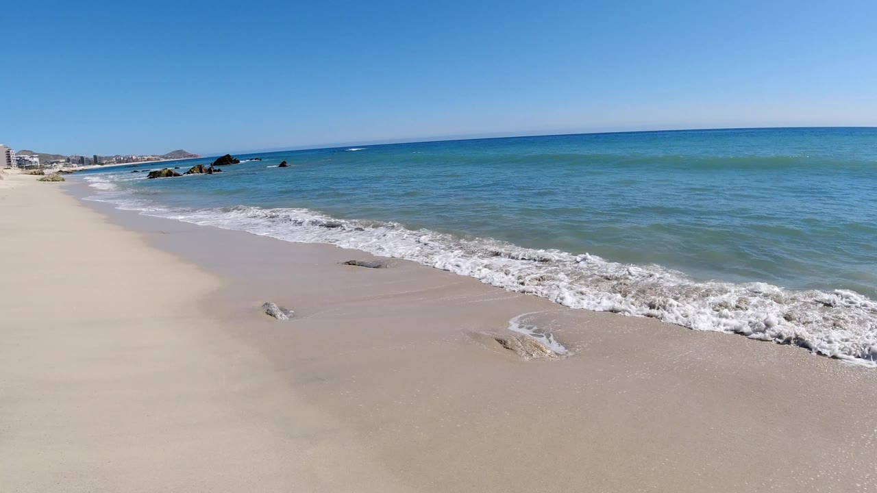 Shoreline curving toward distant buildings, rocks, and blue ocean.