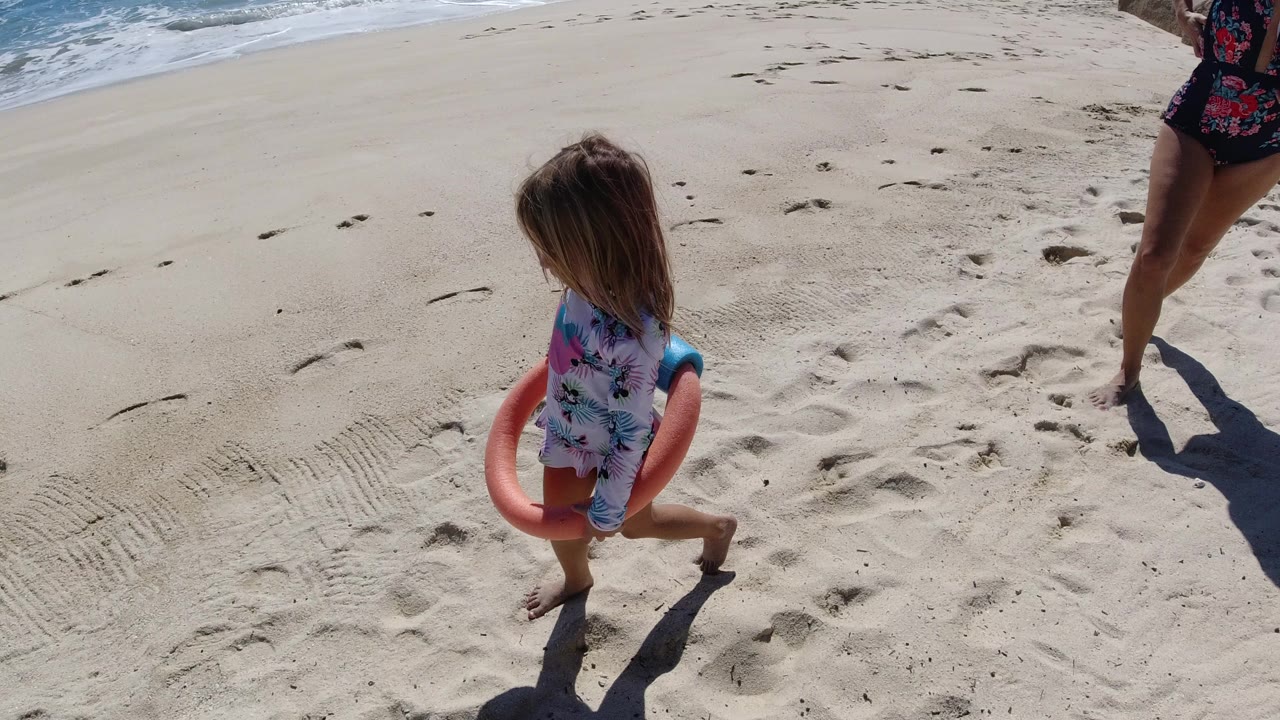 Young girl in a patterned swimsuit walks barefoot on a Los Cabos beach carrying a pink flotation toy, with ocean water at the upper left and adult legs partly visible at the right edge.