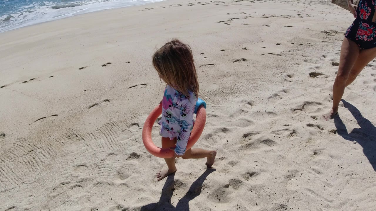Young girl continues walking across the Los Cabos sand with a pink-and-blue toy around her arm, while partial adult legs and long shadows remain near the top right.