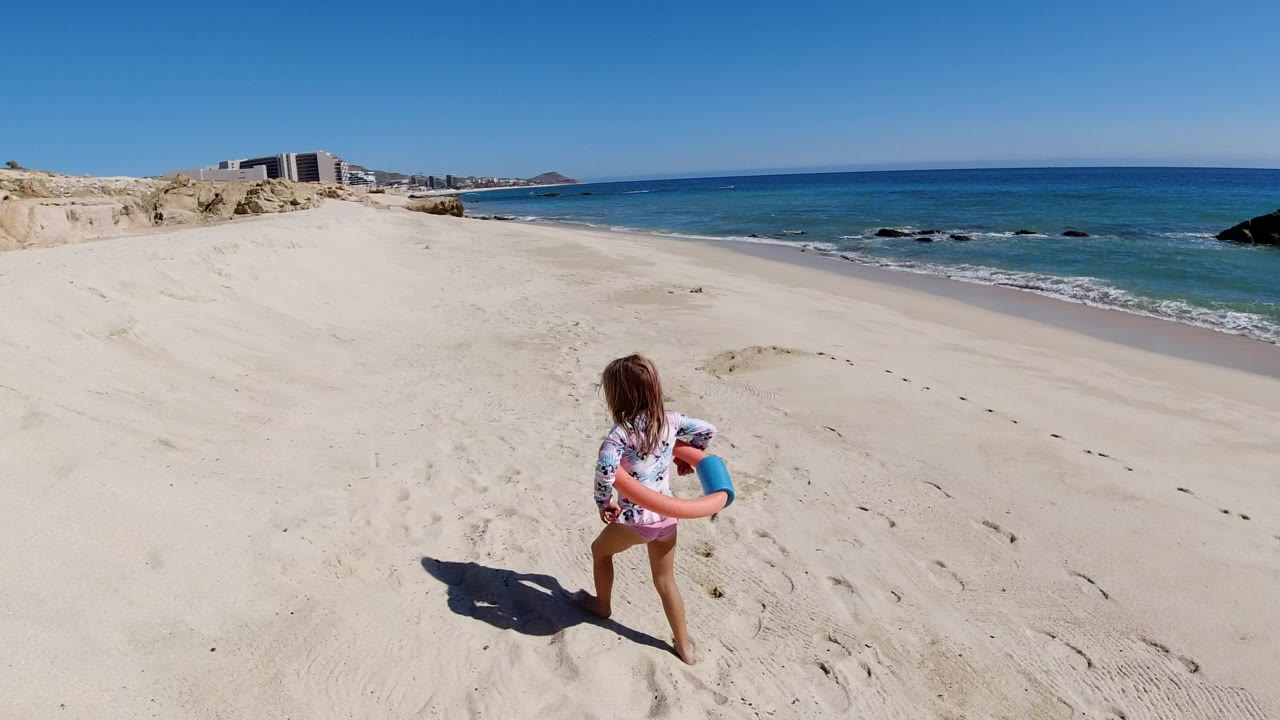 A young girl in Los Cabos carries an orange and blue swim float across pale sand, with blue water, low surf, rocky dunes, and distant buildings behind her.