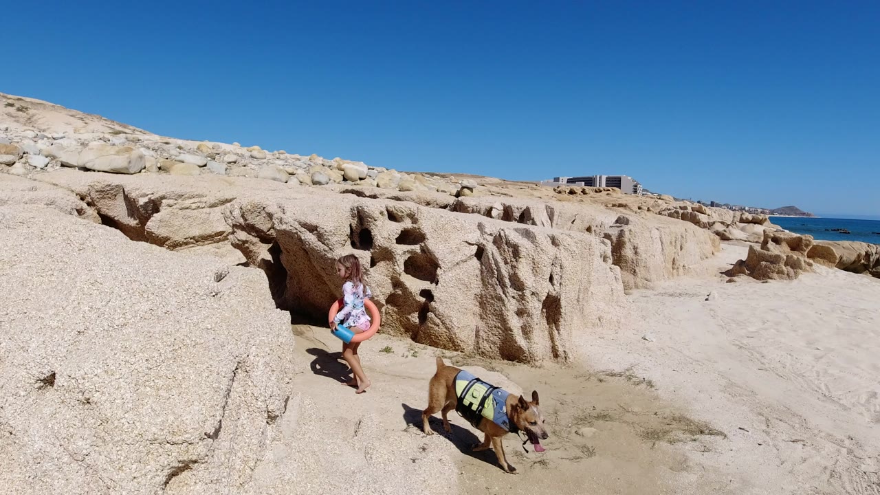 A young girl in Los Cabos, Mexico carries an orange water tube beside eroded pale rocks while a dog in a vest stands on the sandy foreground.