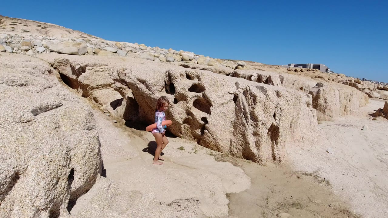Child with swim ring walking beside eroded sand rocks under blue sky.