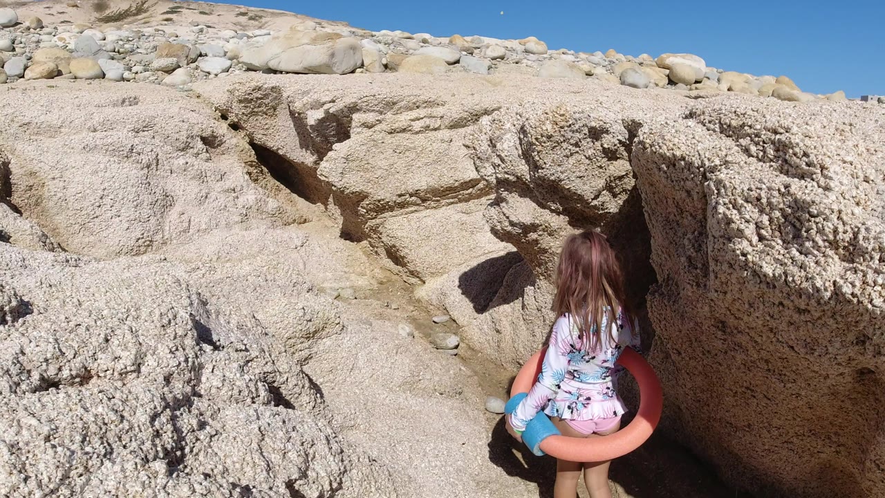 The child faces away from the camera with a pink swim tube around the waist, framed by rough beige rocks and bright blue sky in Los Cabos, Mexico.