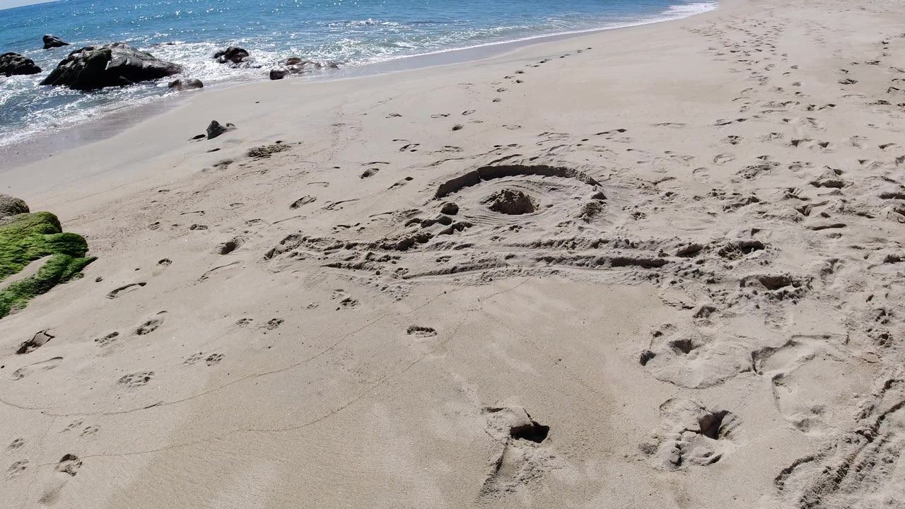 Empty sandy beach with rocks, surf, and a circular mark in the sand.