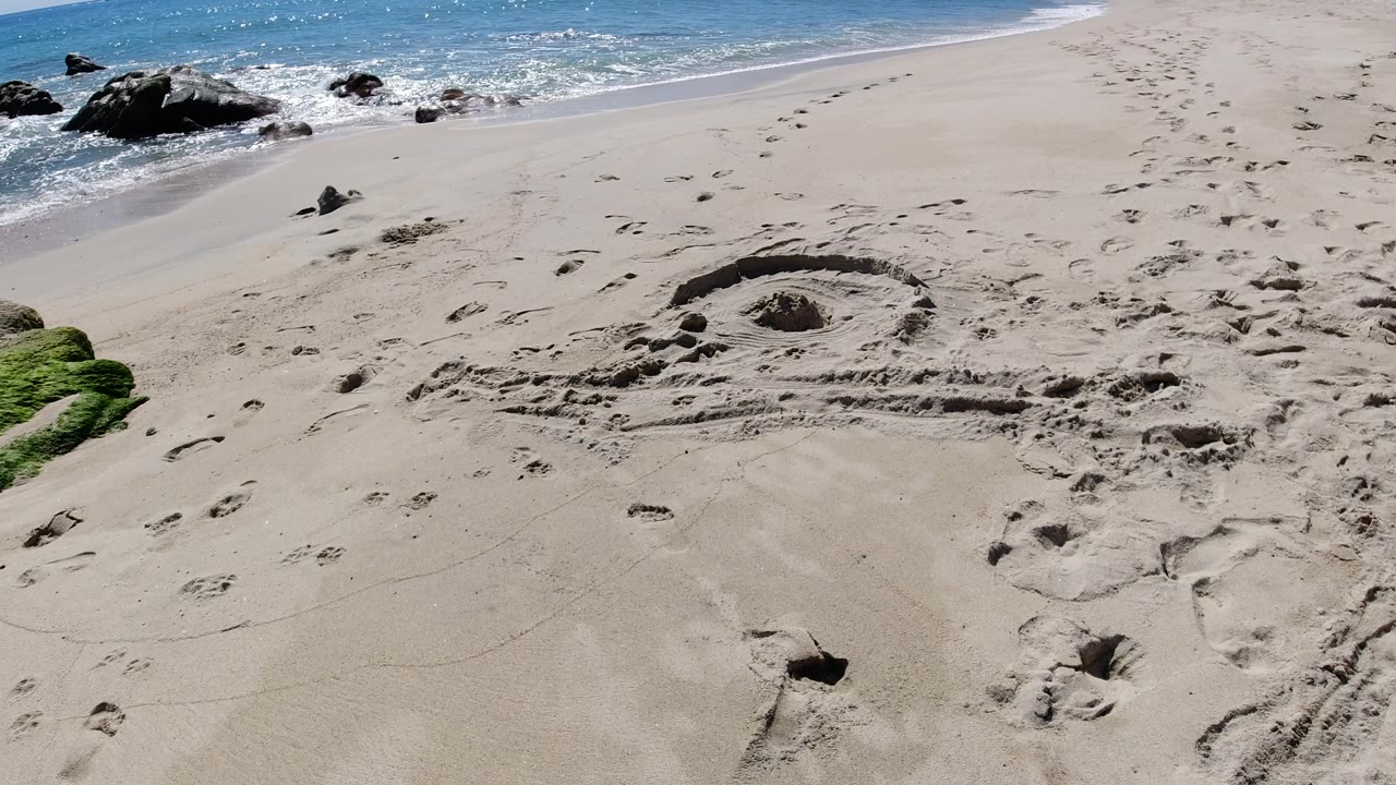 Los Cabos shoreline frame with the sand drawing centered lower in view, footprints crossing the beach, black rocks at the waterline, and no visible people.