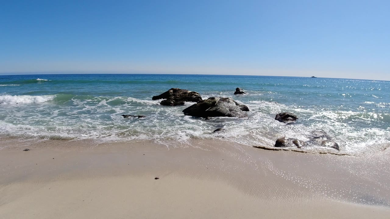 Los Cabos beach shoreline with pale sand, foamy surf around dark rocks, blue ocean, a clear sky, and no visible people at the start of the clip.