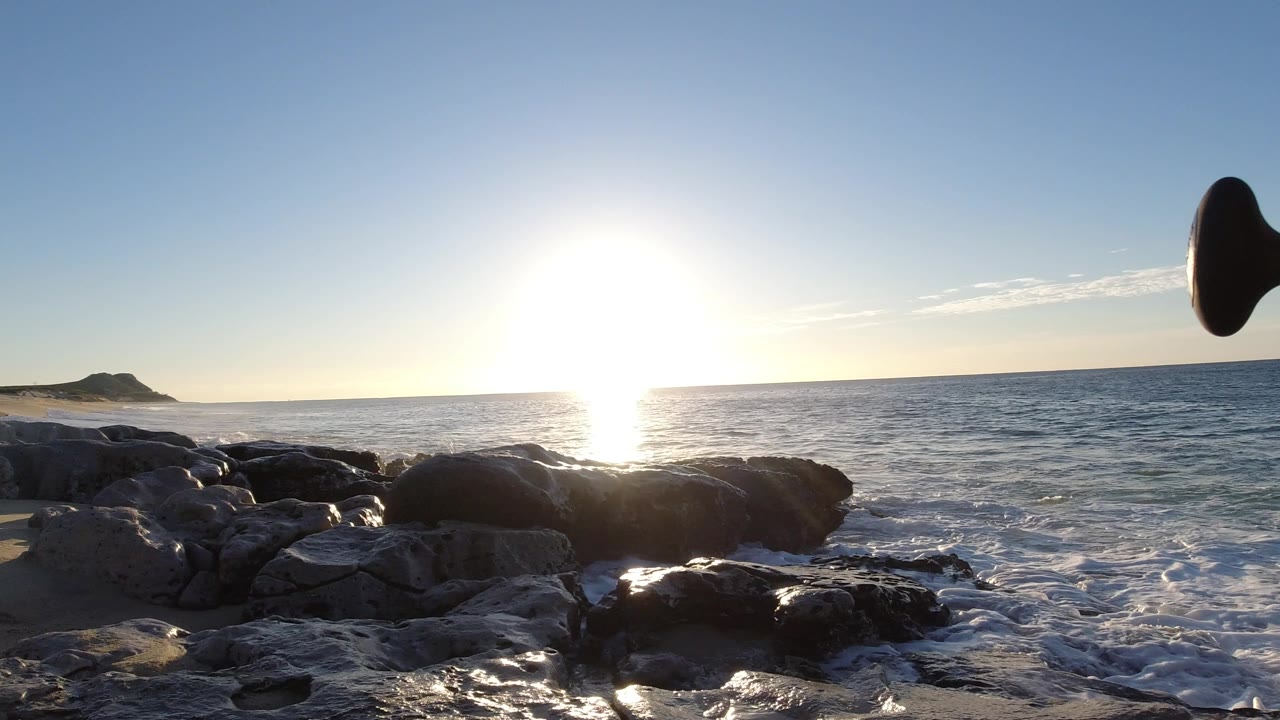 Opening view of the Los Cabos coastline with bright sun glare on the water, dark rocks in the foreground, a distant headland at left, and no visible people.