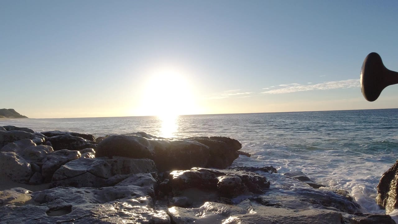 Sunlit ocean with rocks and a dark curved object at frame edge.