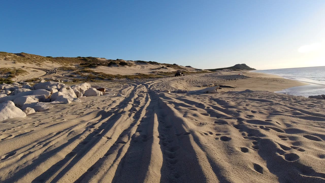 Wide ground-level view of a Los Cabos beach with rutted sand in the foreground, pale rocks at left, low dunes ahead, ocean at right, and no visible people.
