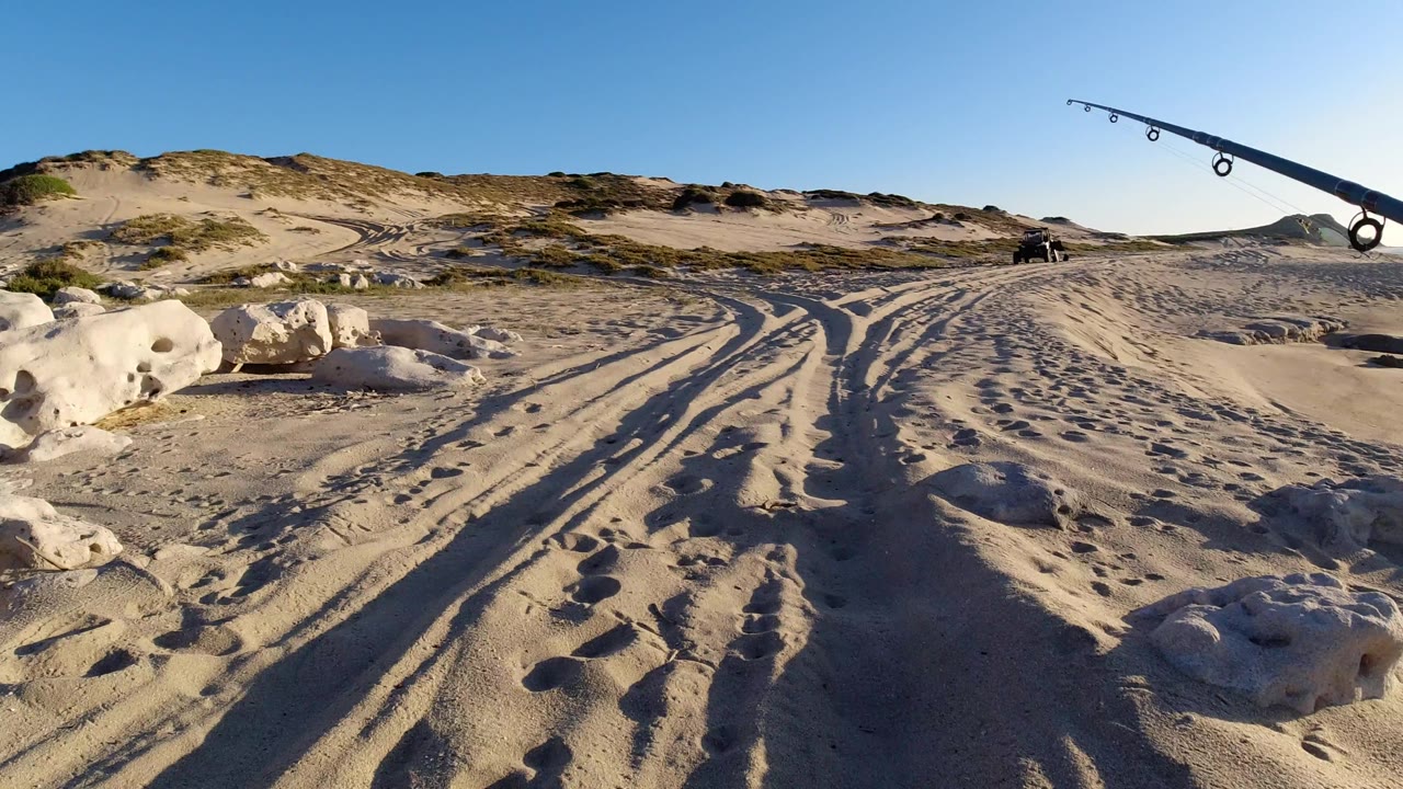 Los Cabos beach frame with sand tracks leading past rounded rocks toward low dunes, while a fishing rod crosses the upper right of the frame.