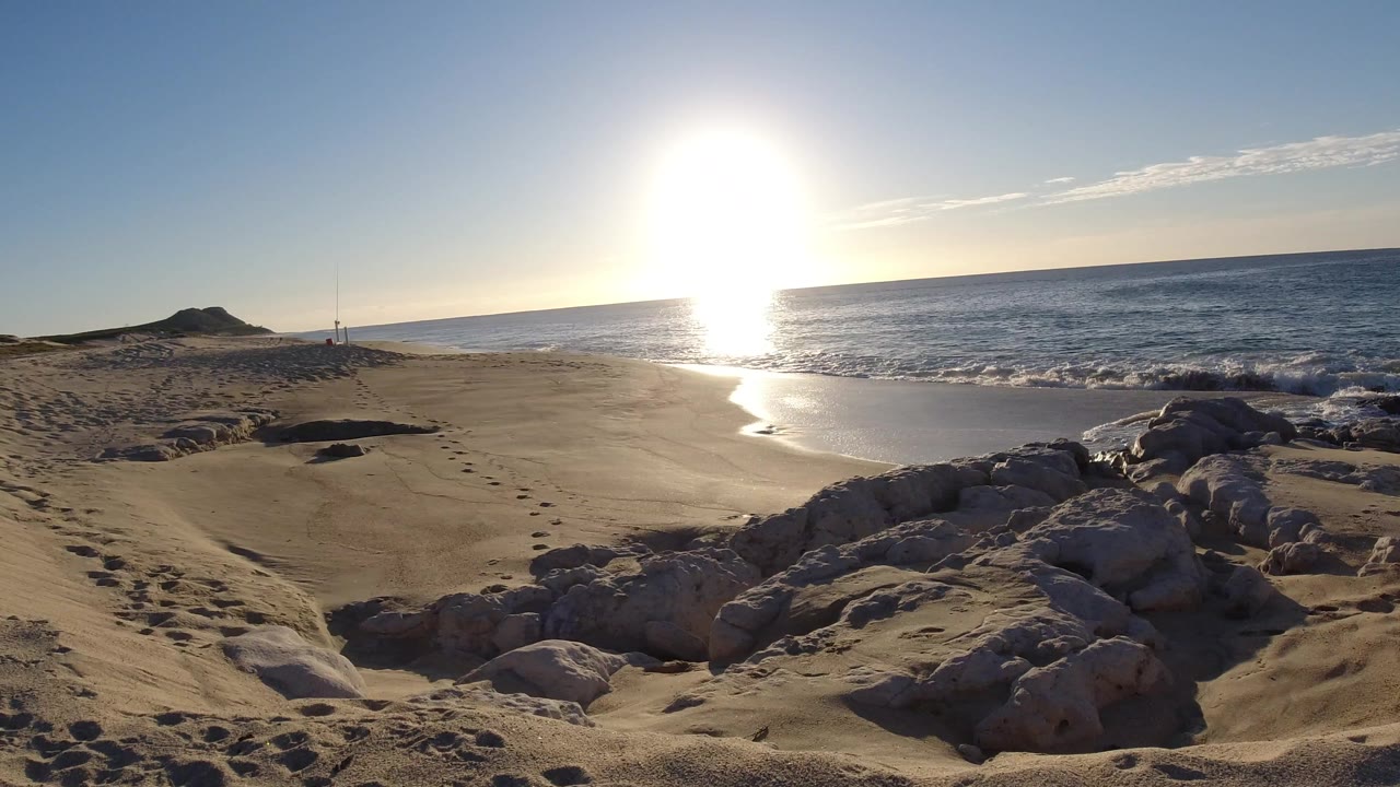 Los Cabos shoreline seen across pale foreground rocks, with a curved wet-sand edge, ocean horizon, bright glare, and no visible people.