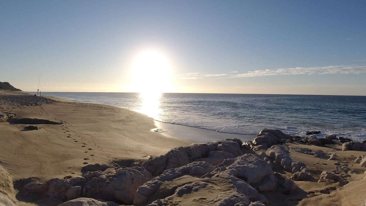 Los Cabos beach view at sunset with tan rocks in the foreground, a sandy shore, ocean waves, a bright sun reflection, and no visible people.