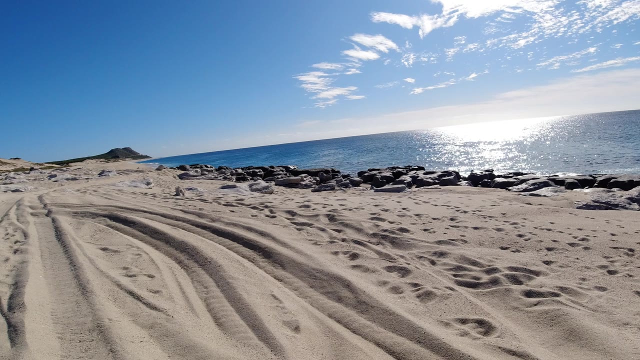 Los Cabos beach view with tire tracks and footprints in pale sand, dark shoreline rocks, bright ocean glare, a small rocky point at left, and no visible people.