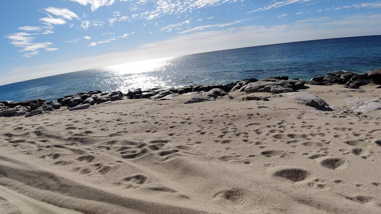 Sandy beach with footprints, rocks, ocean glare, and blue sky.
