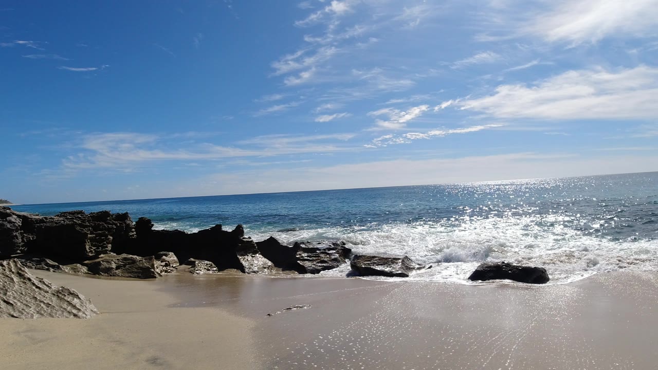 Los Cabos shoreline with foamy water spreading around low black rocks, wet sand in front, open blue ocean behind, and no visible people.