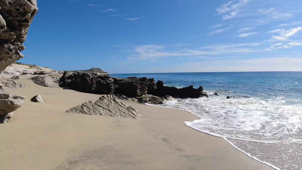 Empty sandy beach with surf, dark rocks, blue ocean, and a rock overhang.
