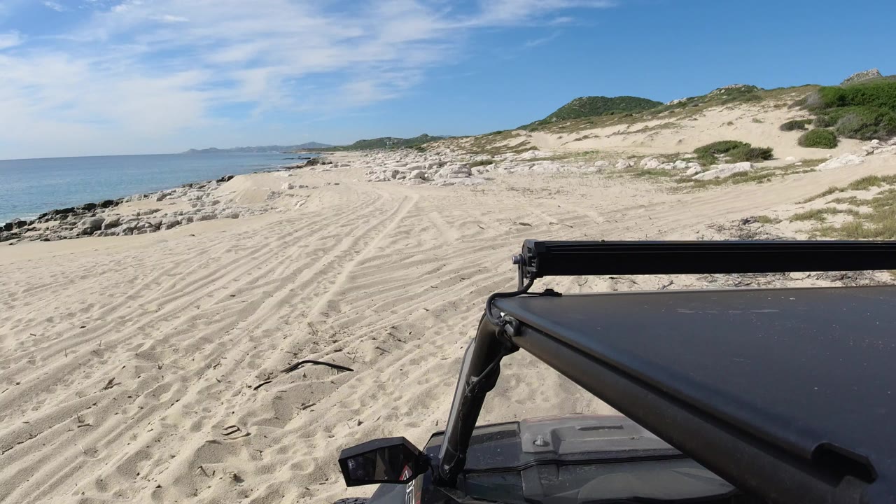 Opening frame of a Los Cabos beach area with a black Wrangler roof and light bar in the foreground, tire-marked sand, ocean at left, scrubby dunes at right, and no visible people.