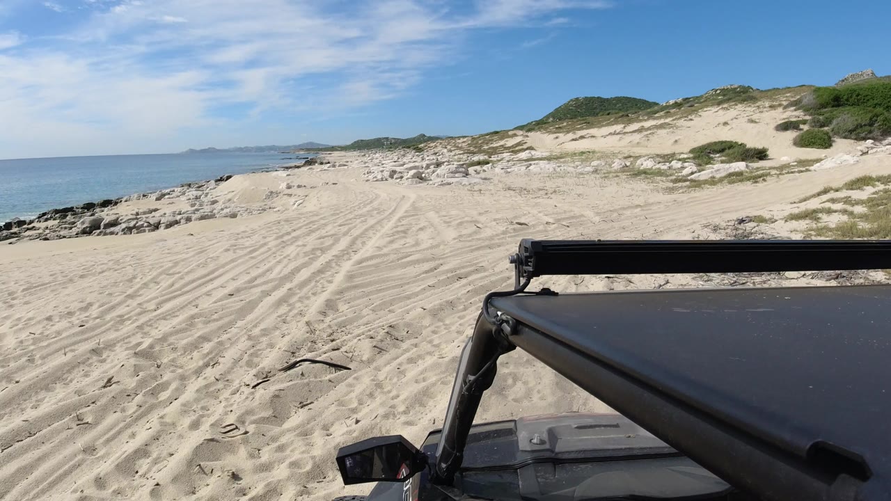Los Cabos beach access view with the Wrangler roof filling the lower right, rutted sand leading toward rocks, blue ocean on the left, low green dunes, and no visible people.