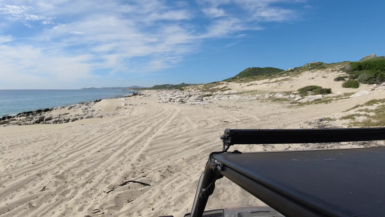 Opening view of a Los Cabos sandy beach with tire tracks, blue ocean on the left, dune scrub on the right, a dark foreground edge, and no visible people.