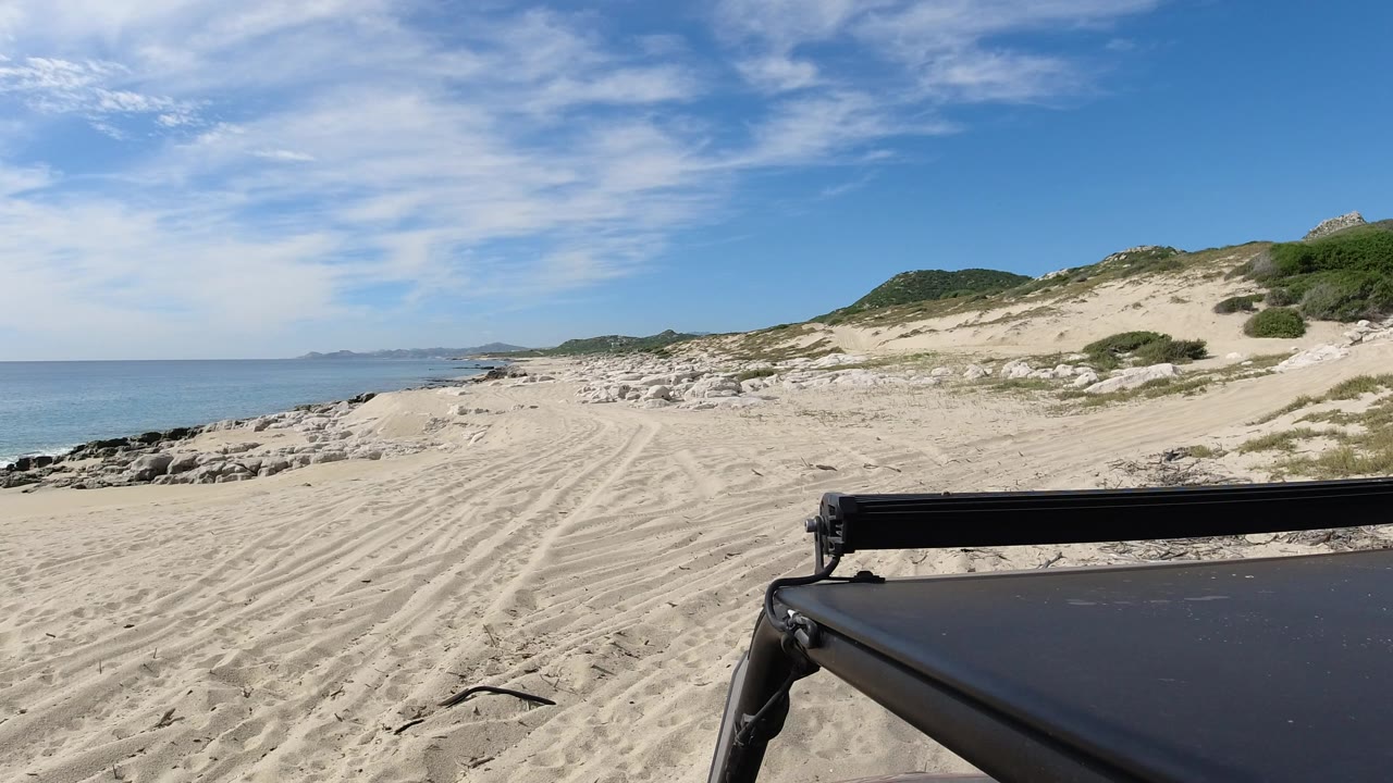 Sandy beach with ocean, dunes, and a dark vehicle edge in the foreground.