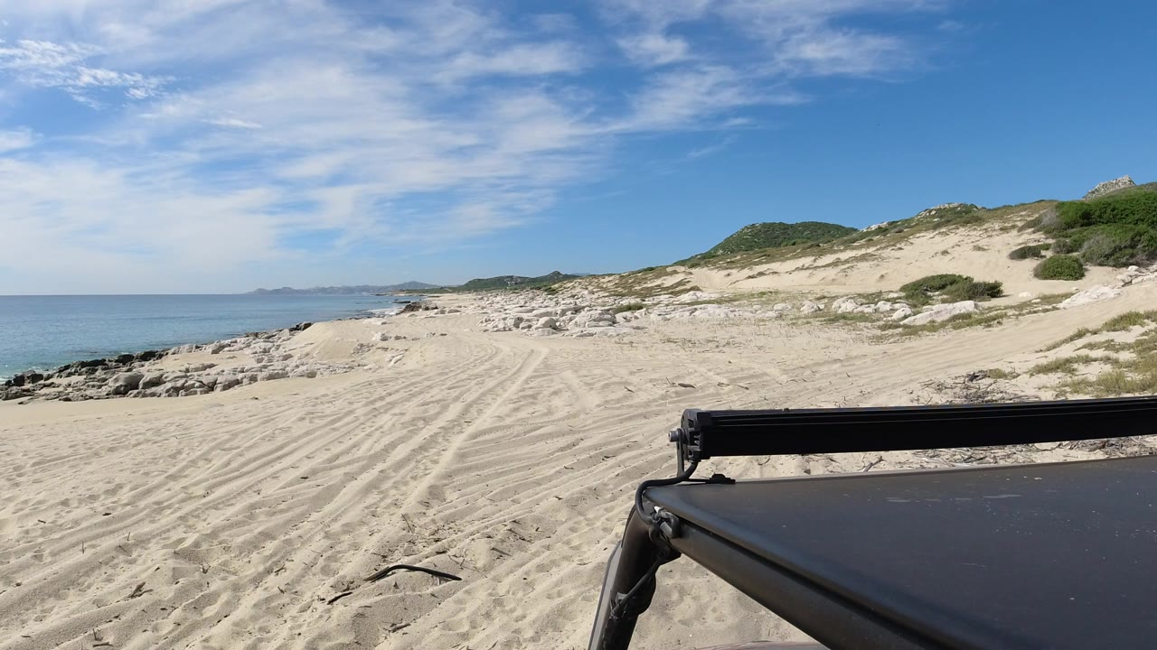 Opening frame of a Los Cabos beach view with pale sand tracks, blue ocean at left, low dunes at right, a black vehicle edge in the foreground, and no visible people.