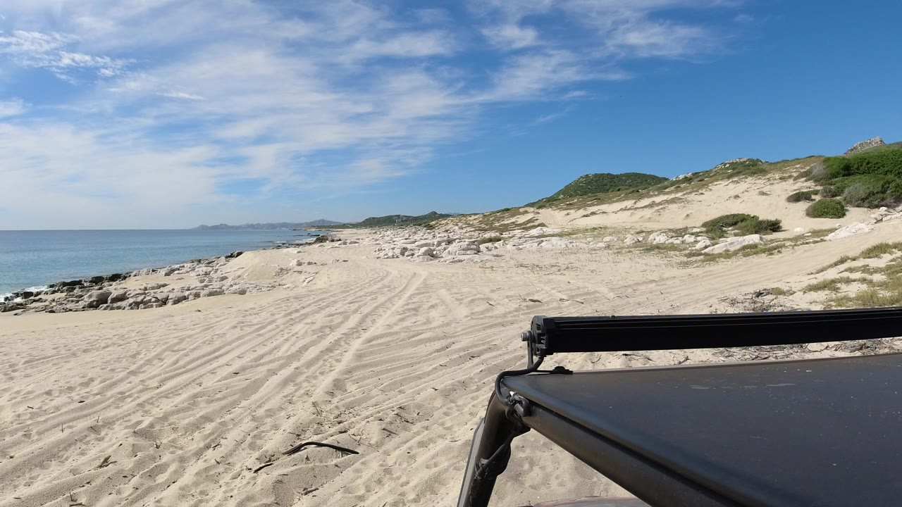 Vehicle-mounted view along a sandy Los Cabos beach, with the Jeep roof and black bar in the lower foreground, ocean on the left, dunes on the right, and no visible people.