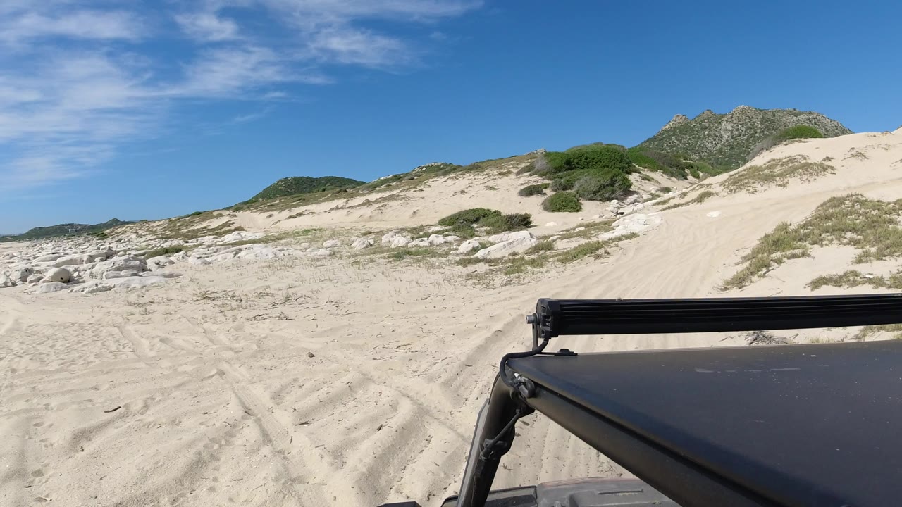 The Jeep-mounted camera looks across pale sand in Los Cabos, Mexico, with tire ruts leading toward scrub-covered dunes, white rocks, distant hills, and no visible people.