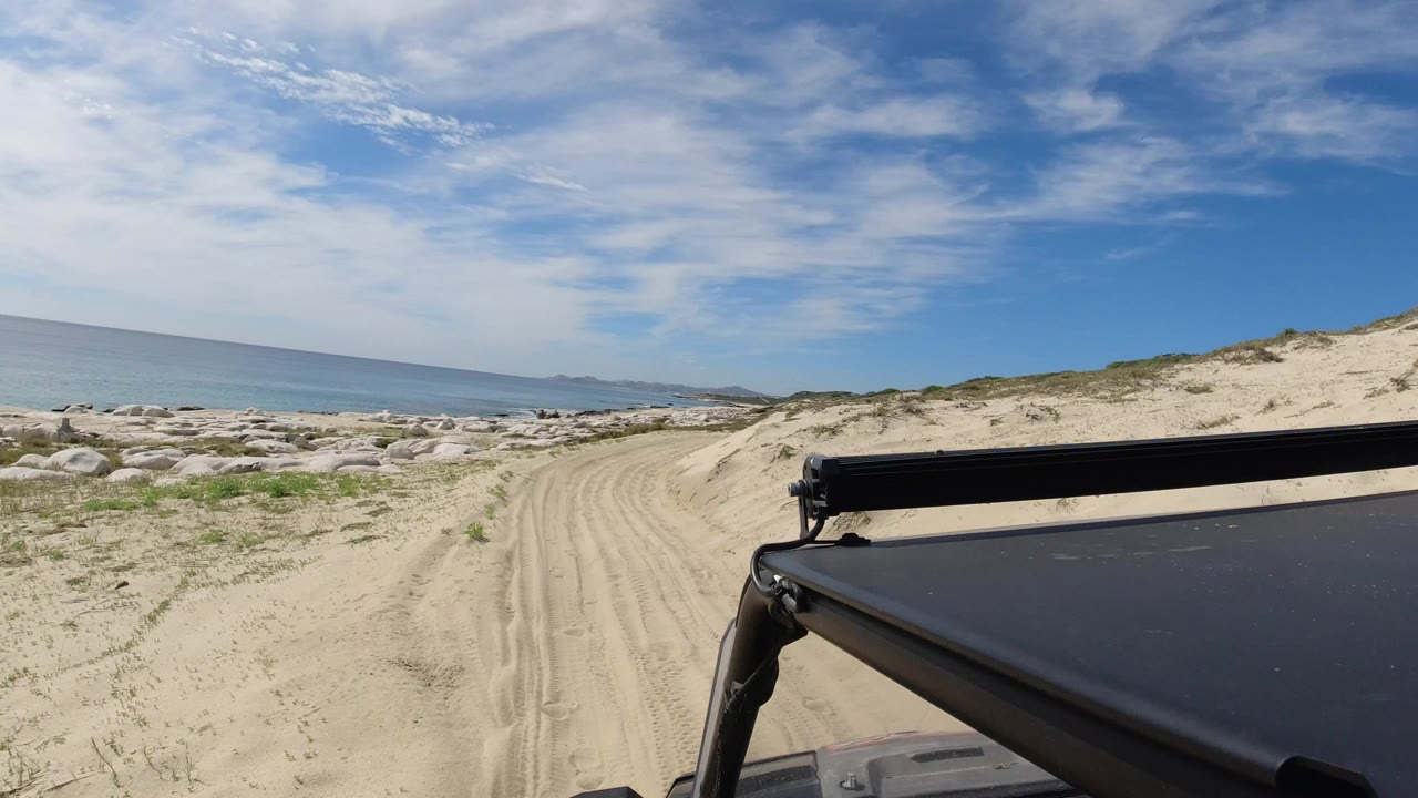 Sandy beach road beside ocean and dunes with vehicle roof in foreground.