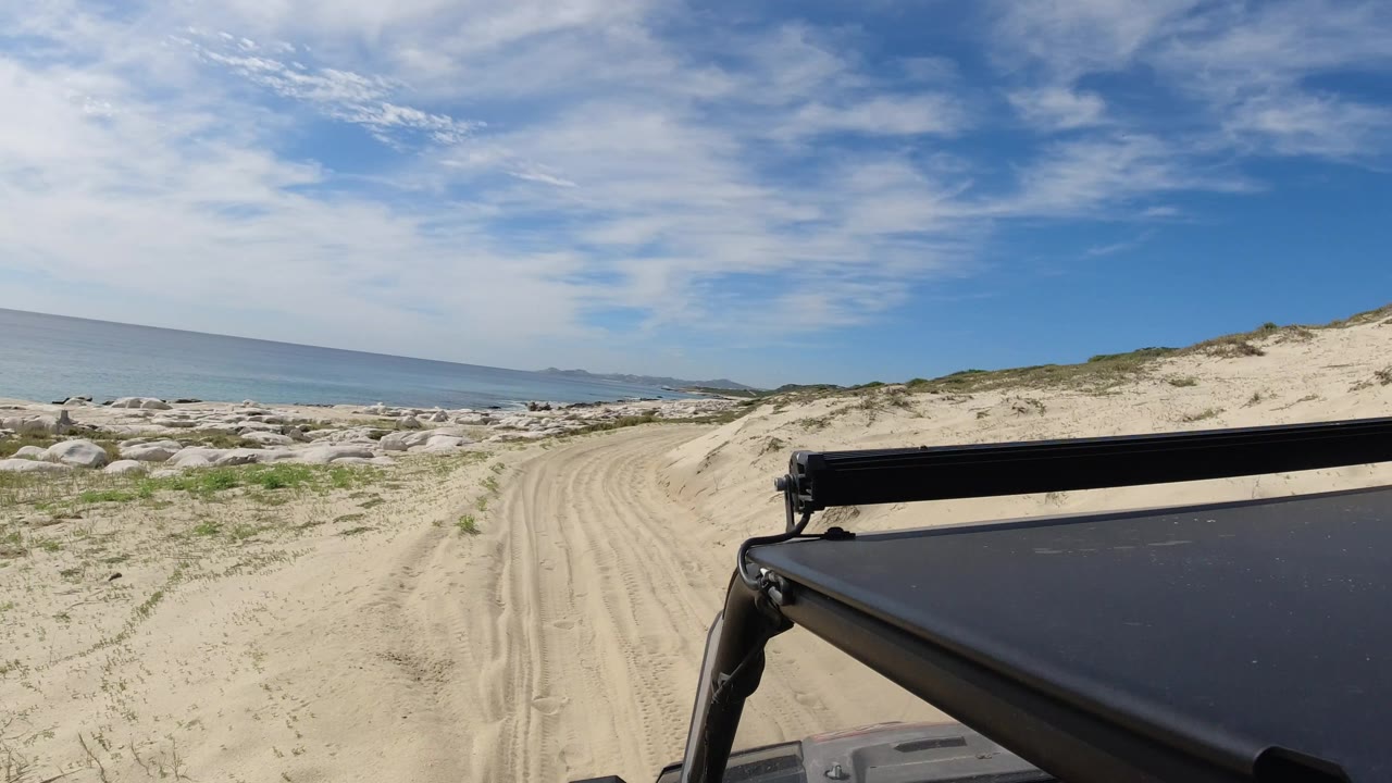 Los Cabos beach road continues ahead with ocean on the left, scattered shoreline rocks, low dune vegetation, a blue clouded sky, and no visible people.