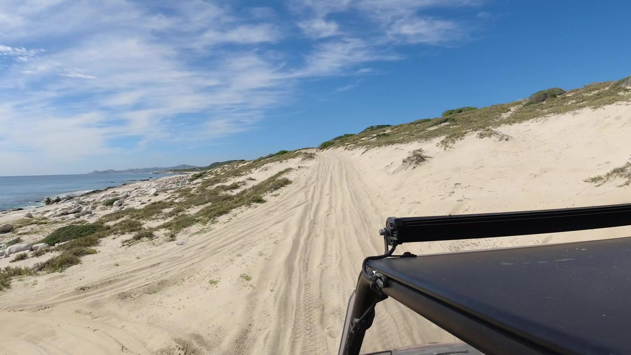 A sandy vehicle track runs through low dune plants beside ocean water in Los Cabos, Mexico, with a dark vehicle roof and rack at the lower right and no visible people.