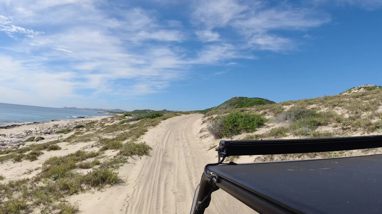 Los Cabos, Mexico road-cam view of a sandy track curving through low dunes beside the ocean, with a black vehicle roof in the foreground and no visible people.