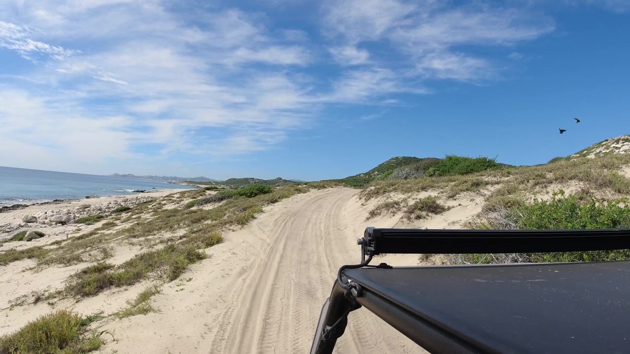 Sandy road beside dunes with ocean and distant hills under blue sky.