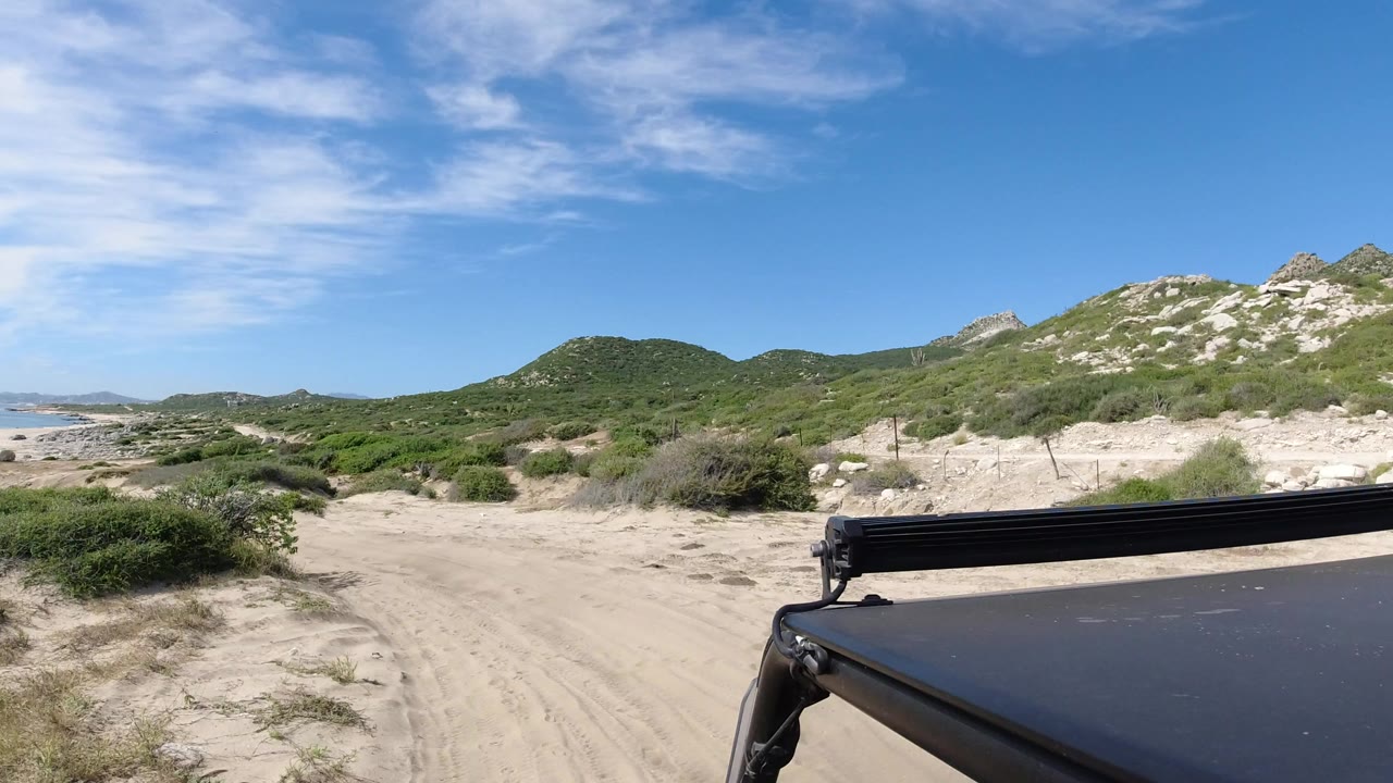 Opening view of a sandy road through coastal scrub in Los Cabos, Mexico, with the ocean at left, green hills ahead, a dark vehicle roof edge at lower right, and no visible people.