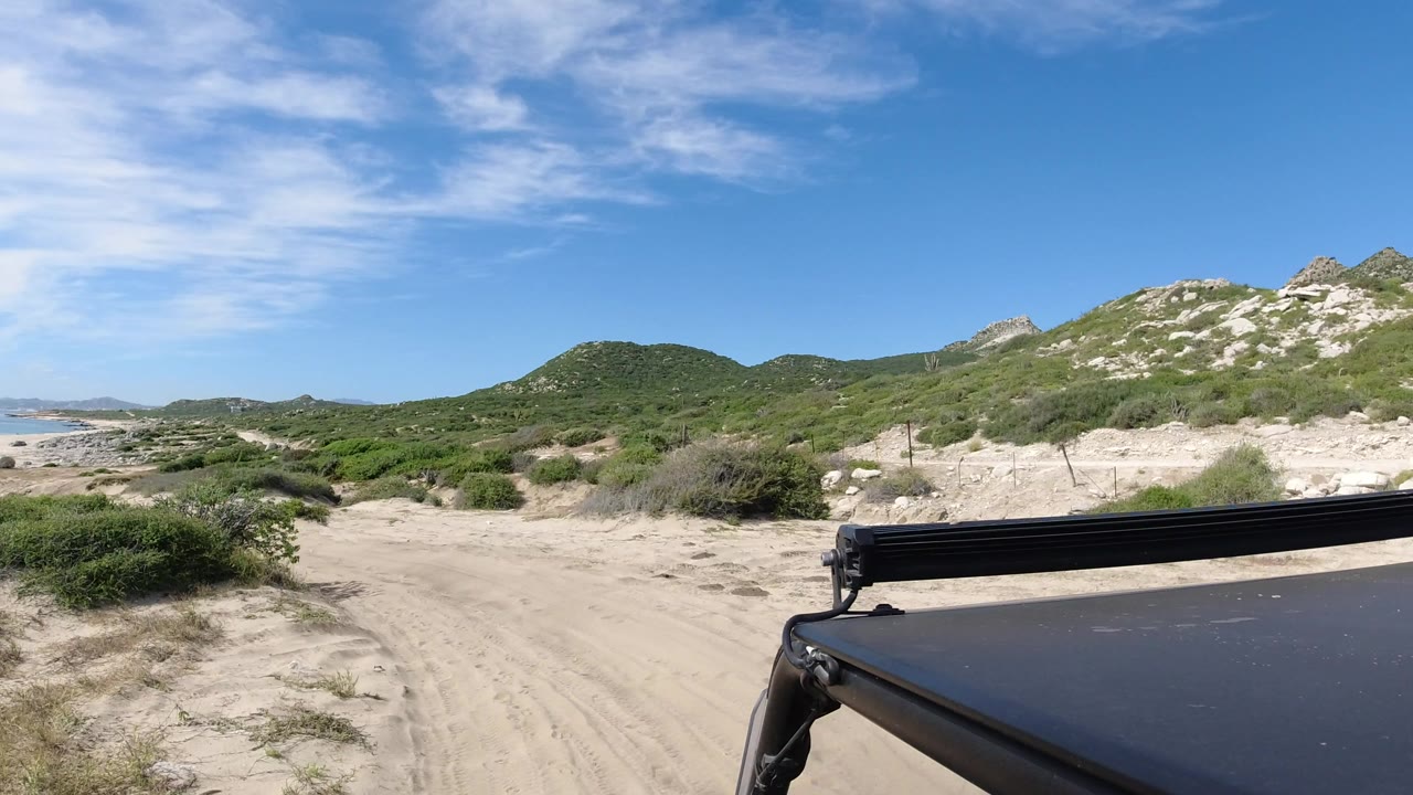 A sandy track curves through low vegetation in Los Cabos, Mexico, with pale rocky hillside at right, distant water at left, the vehicle roof in the foreground, and no visible people.