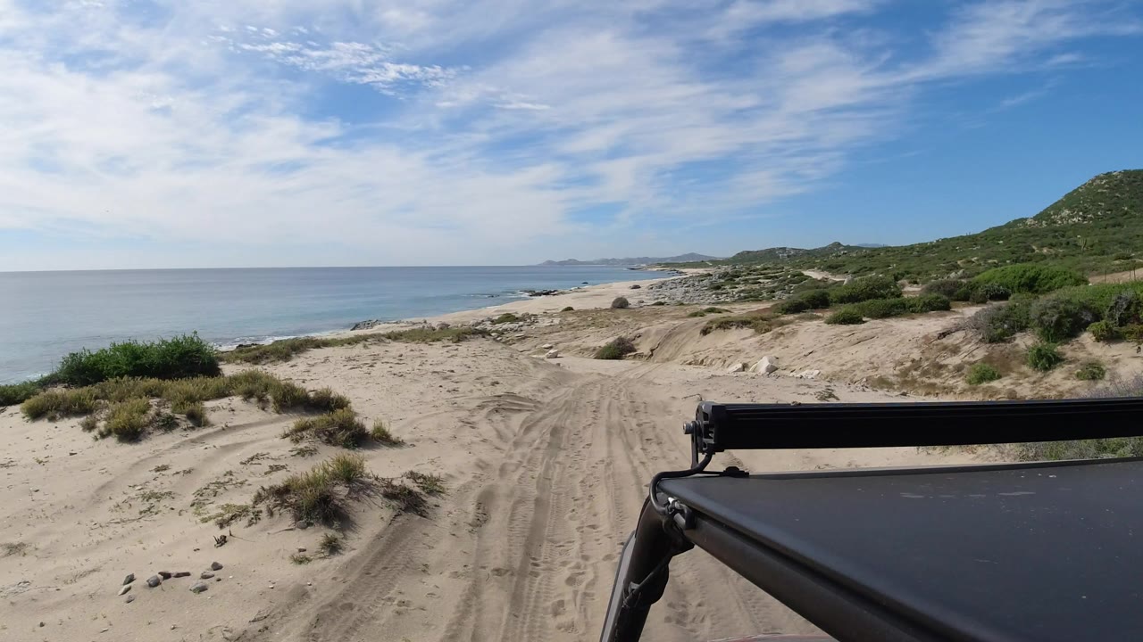 Sandy beach track beside blue water and green hills with a dark vehicle edge in front.