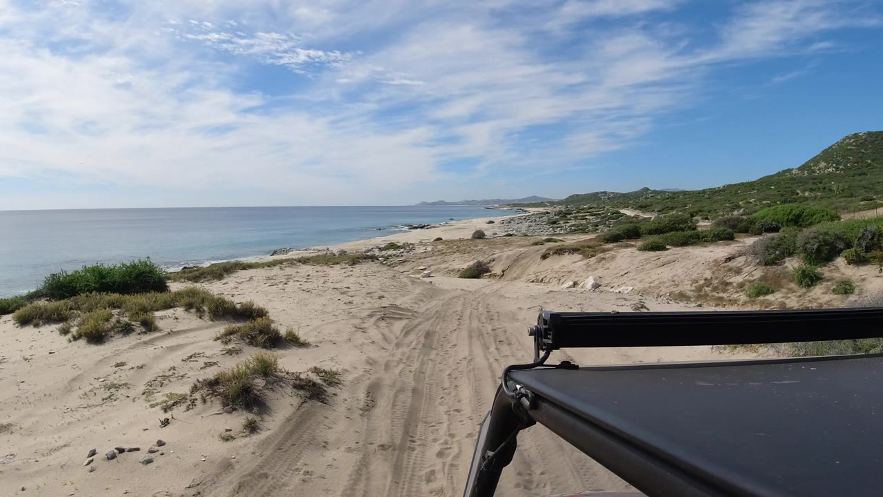 A Los Cabos sandy track continues along the coast, with calm water to the left, scrub-covered dunes ahead, a green hill on the right, vehicle hardware in the foreground, and no visible people.