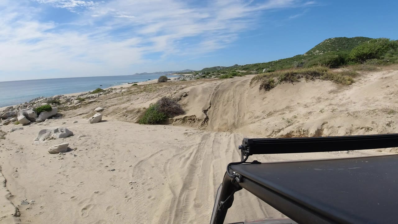 Los Cabos, Mexico sand road continues past low shrubs and beach rocks, with the ocean on the left horizon, hills ahead, vehicle roof at bottom right, and no visible people.