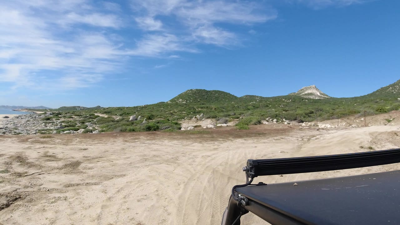 Vehicle-level view in Los Cabos, Mexico of a sandy track facing low green hills and a pale rocky ridge under blue sky, with no visible people.