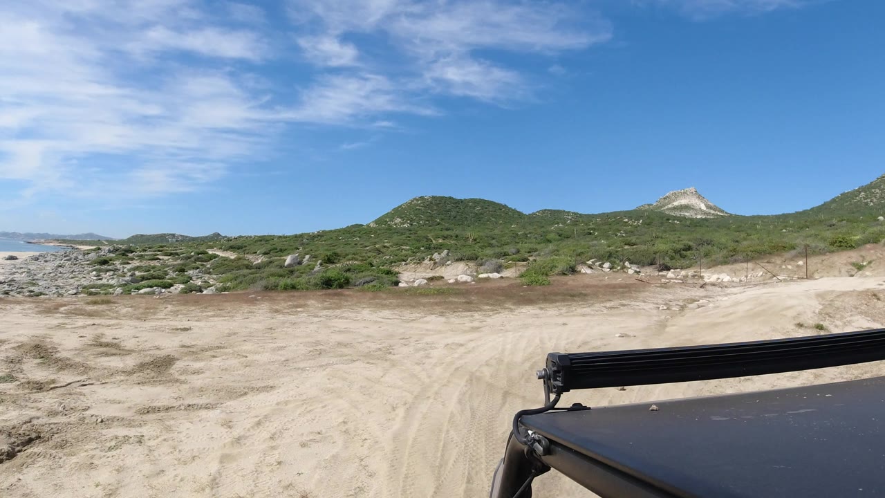 Los Cabos coastal desert view with sand tracks, scattered scrub, low hills, a light rock outcrop, and black vehicle hardware crossing the lower right, with no visible people.