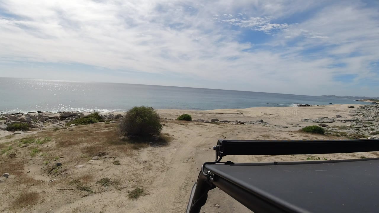Opening frame from Los Cabos shows a sandy coastal track, low brush, scattered rocks, blue ocean, bright clouded sky, and no visible people.