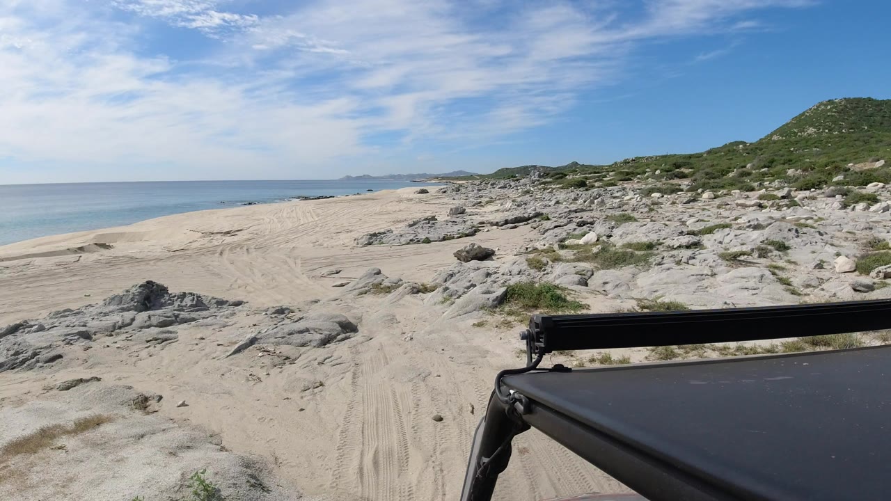 Beach sand, rocks, scrub, calm sea, and vehicle roof edge under a bright sky.