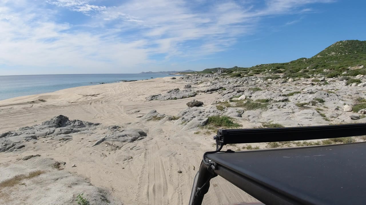 A slightly advanced Los Cabos beach view shows the vehicle roof and bar at lower right, sand tracks leading ahead, rock outcrops, scrub, calm water, and no visible people.
