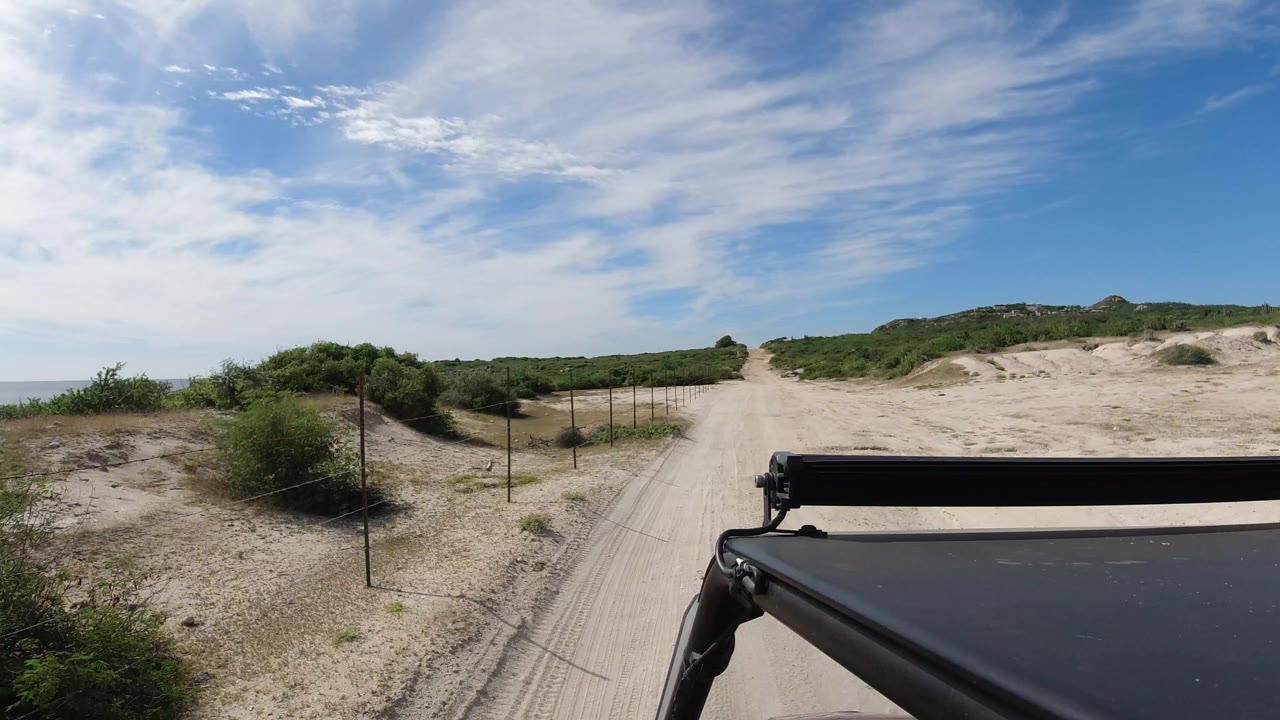 Start of the Los Cabos, Mexico clip showing a sandy road between low green brush and wire fence posts, with part of the Wrangler roof or frame at lower right and no visible people.