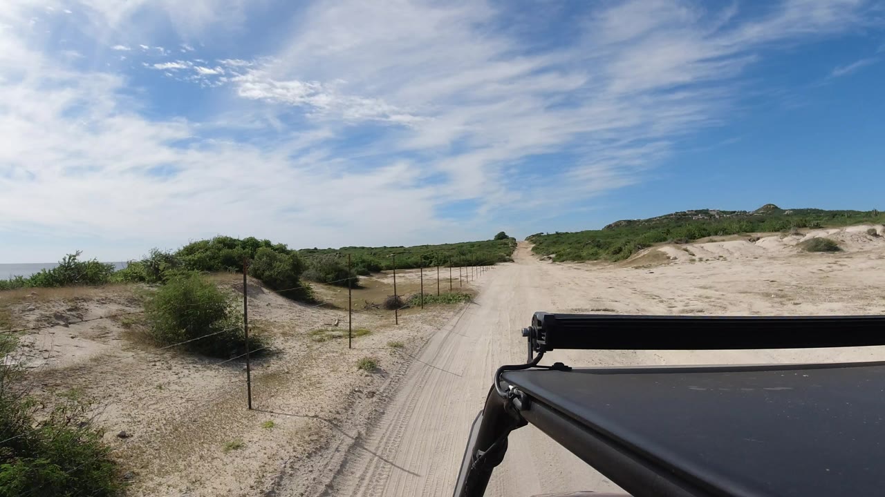 One second into the Los Cabos, Mexico footage, the sandy road runs ahead beside thin fence posts and green scrub, with the dark Wrangler roof edge fixed along the lower right and no visible people.
